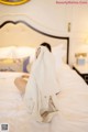 A woman sitting on a bed covered in a white robe.