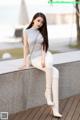 A woman sitting on a concrete wall wearing white pants and boots.